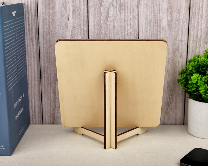 Puede incluir: Un marco de fotos de madera con un acabado natural, sostenido por un caballete de madera. El marco es cuadrado con esquinas redondeadas. El soporte tiene una base triangular y un soporte vertical. Un libro azul y una peque&ntilde;a planta en maceta est&aacute;n en el fondo.