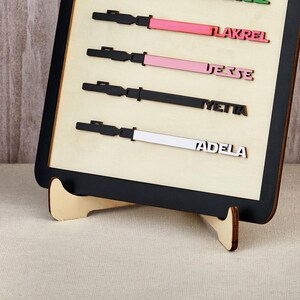 Puede incluir: Un letrero de madera con marco negro y fondo beige claro. El letrero presenta cuatro dise&ntilde;os de madera cortados con l&aacute;ser con los nombres: LAKREL, JESSE, YETTA y ADELA. Cada dise&ntilde;o tiene un mango negro y una hoja de color: rosa, rosa, negro y blanco.