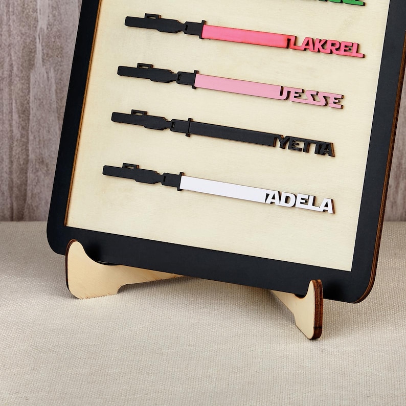 Puede incluir: Un letrero de madera con marco negro y fondo beige claro. El letrero presenta cuatro dise&ntilde;os de madera cortados con l&aacute;ser con los nombres: LAKREL, JESSE, YETTA y ADELA. Cada dise&ntilde;o tiene un mango negro y una hoja de color: rosa, rosa, negro y blanco.