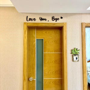 May include: A wooden door with a light brown frame and a frosted glass panel. Above the door, the phrase "Love You, Bye" is written in black script. A small decorative plant is mounted on the wall to the right of the door.