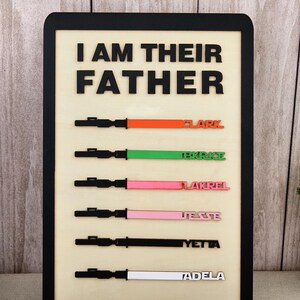 Puede incluir: Letrero de madera con el texto "I AM THEIR FATHER" en letras negras. Debajo, seis dise&ntilde;os de sables de luz en varios colores, cada uno con un nombre: Clark, Bkrice, Lakrel, Jesse, Yetta y Adela.