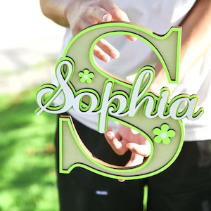 May include: A wooden decorative sign in the shape of the letter "S" with the name "Sophia" in white script. The letter and name are outlined in bright green, with small flower accents. The sign is held up by a person.