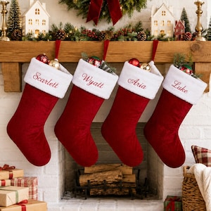 Könnte beinhalten: Vier tiefrote Weihnachtsstrümpfe aus Samt mit weißem Kunstfellbesatz, jeweils mit einem in roter Schreibschrift eingestickten Namen: Scarlett, Wyatt, Aria und Luke. Jeder Strumpf hat eine rote Aufhängeschlaufe und enthält kleine Dekorationen.