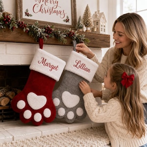 May include: Two paw-shaped Christmas stockings with white sherpa cuffs. One stocking is red with the name Margot embroidered in red, and the other is gray with the name Lillian embroidered in red. Both feature white sherpa paw pads.