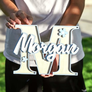 May include: A wooden letter "M" with the name "Morgan" in cursive script. The letter is light beige with a light blue outline and accents, including small flower details. The layered design creates a 3D effect.