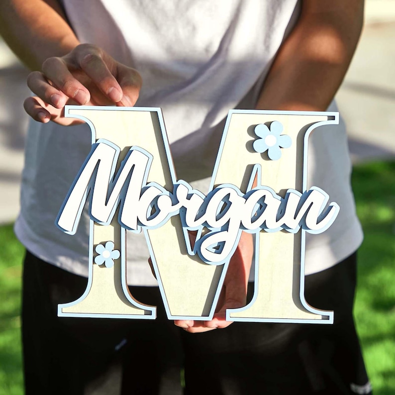 May include: A wooden letter "M" with the name "Morgan" in cursive script. The letter is light beige with a light blue outline and accents, including small flower details. The layered design creates a 3D effect.
