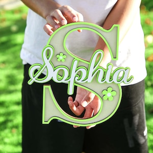 May include: A wooden sign in the shape of the letter "S" with the name "Sophia" in white script. The sign has a light green outline and small flower accents. The sign is held in front of a blurred green background.