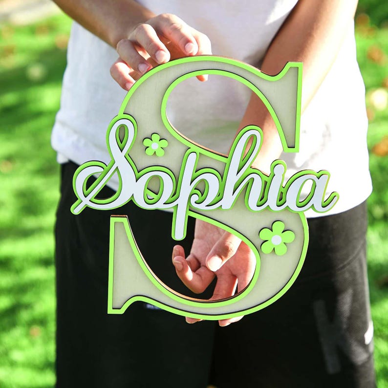 May include: A wooden sign in the shape of the letter "S" with the name "Sophia" in white script. The sign has a light green outline and small flower accents. The sign is held in front of a blurred green background.