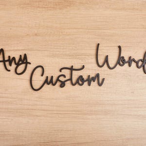 Puede incluir: Recortes de letras en cursiva negra que forman las palabras Any Custom Words. Las letras son piezas individuales dispuestas en tres líneas separadas, con un acabado mate suave y un diseño de tipografía fluida y conectada.