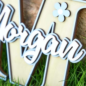 May include: Wooden name sign with the name "Morgan" in white script lettering, layered over a light blue and brown background. A light blue flower is attached to the sign. The sign is placed on a bed of green grass.