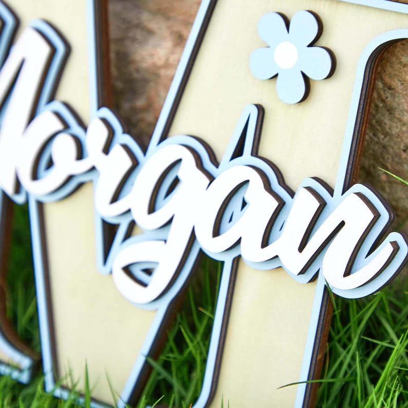 May include: Wooden name sign with the name "Morgan" in white script lettering, layered over a light blue and brown background. A light blue flower is attached to the sign. The sign is placed on a bed of green grass.