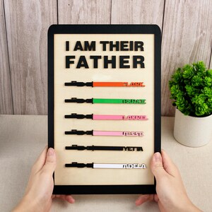 Puede incluir: Cartel de madera con la frase "I AM THEIR FATHER" en letras negras. Debajo, dise&ntilde;os de sables de luz coloridos con nombres: Flark, Tekrise, Lakrel, Jesse, Yetta y Tadela. El cartel est&aacute; enmarcado en negro.
