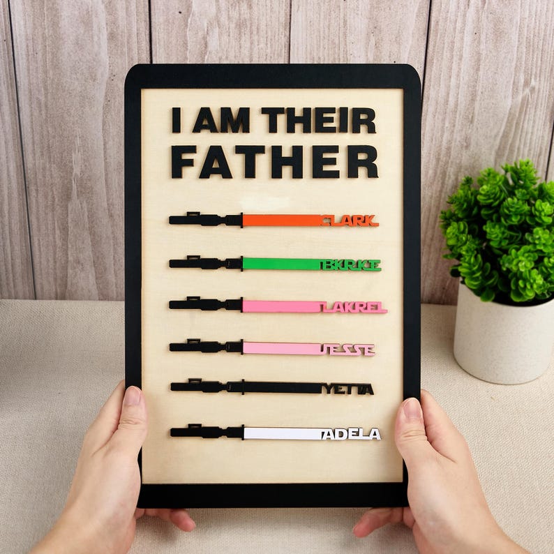 Puede incluir: Cartel de madera con la frase "I AM THEIR FATHER" en letras negras. Debajo, dise&ntilde;os de sables de luz coloridos con nombres: Flark, Tekrise, Lakrel, Jesse, Yetta y Tadela. El cartel est&aacute; enmarcado en negro.