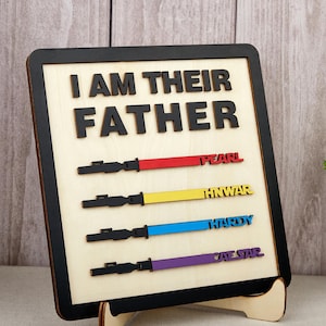 May include: Wooden sign with the phrase "I AM THEIR FATHER" in black letters. Below are four colorful lightsaber designs with names: Pearl, Hnwar, Hardy, and Cate Star. The sign has a black border and is propped up on a wooden stand.