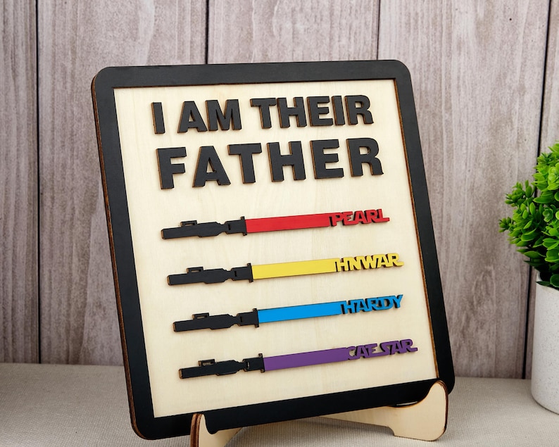 Puede incluir: Letrero de madera con la frase "I AM THEIR FATHER" en letras negras. Debajo, cuatro dise&ntilde;os de sables de luz coloridos con nombres. El letrero tiene un borde negro y est&aacute; apoyado en un soporte de madera.