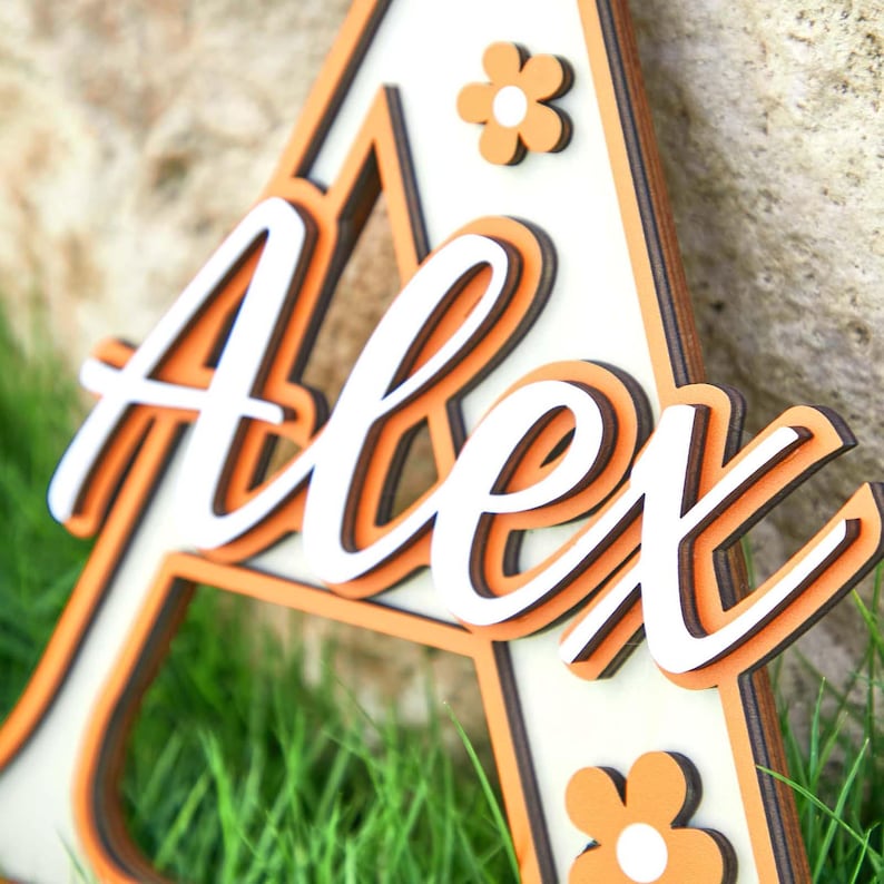 May include: Wooden sign in the shape of the letter "A" with the name "Alex" in white script. The sign has orange and brown accents, with small flower details. The sign is placed on a bed of green grass.