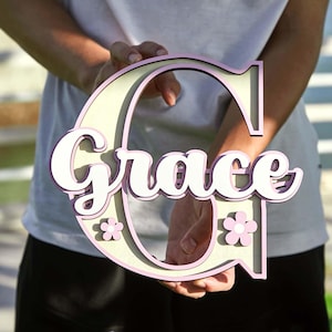 May include: A wooden sign in the shape of the letter "C" with the name "Grace" in white script. The sign has a light beige center with a pink outline and small pink flower accents.