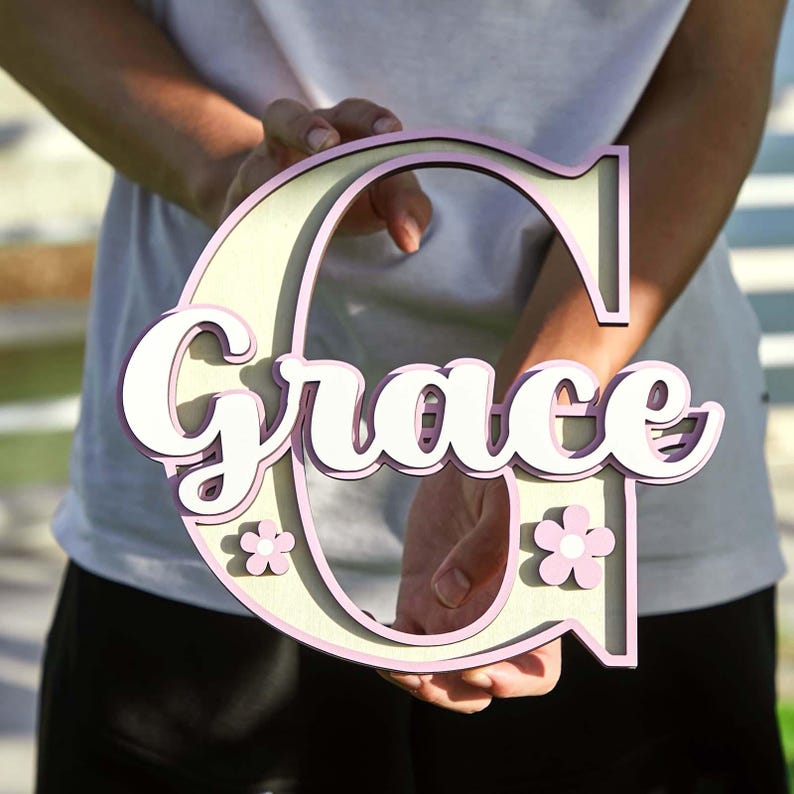 May include: A wooden sign in the shape of the letter "C" with the name "Grace" in white script. The sign has a light beige center with a pink outline and small pink flower accents.