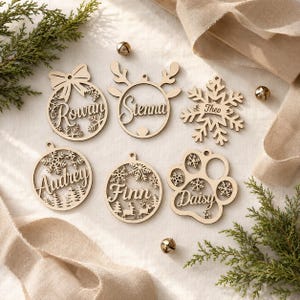 May include: Six laser-cut wooden ornaments featuring names Rowan, Sienna, Theo, Audrey, Finn, and Daisy. The designs include a bow, reindeer antlers, a snowflake, trees, and a paw print, all crafted from light-colored wood with intricate cutouts.