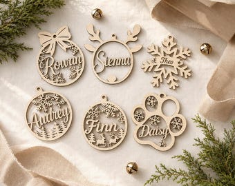 May include: Six light-colored wooden ornaments featuring laser-cut designs and names. The shapes include a bow-topped circle, a reindeer-antler circle, a snowflake, a forest circle, a reindeer circle, and a paw print, personalized with Rowan, Sienna, Theo, Audrey, Finn, and Daisy.