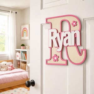 Könnte beinhalten: Ein großes Wand-Buchstabenschild in Form eines R aus hellem Holz mit einem leuchtend pinken Rand. Der Name Ryan ist in weißen Blockbuchstaben in der Mitte des R angebracht und mit zwei kleinen pink-weißen Blumenmotiven verziert.