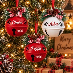 May include: Three round metal jingle bell ornaments hanging from a tree. Two are red and one is white, each featuring a red and white checkered bow, pine needles, and pinecones. The ornaments are personalized with the names Claire, Asher, and Poppy.
