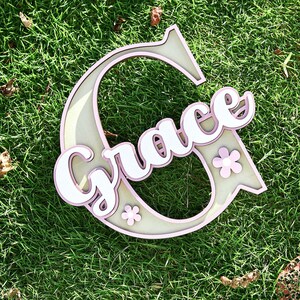 May include: A wooden name sign spelling "Grace" in white script, layered over a light pink and beige letter "G" shape. The design includes small pink flower accents. The sign is placed on a bed of green grass.