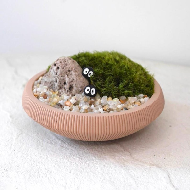 May include: A small, peach-colored, ridged ceramic bowl contains a miniature zen garden. The garden features a small rock, green moss, tiny pebbles, and two black, cartoon-like figures with white eyes. The bowl sits on a white surface.