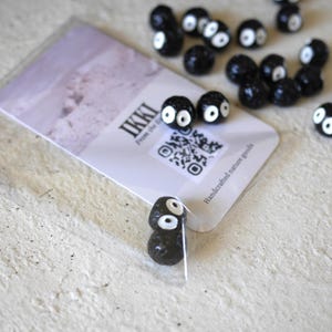 May include: Black, round beads with white eyes are scattered around a card with the word "IKKI" and a QR code. The card also reads "Handcrafted nature goods." The beads have a cartoonish appearance.