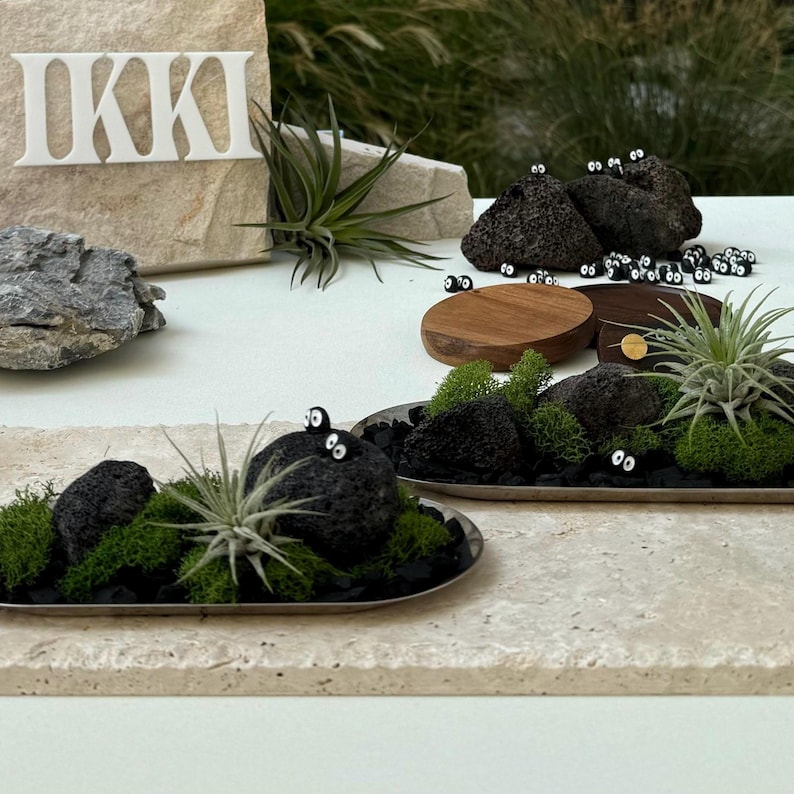 May include: A decorative display featuring small, black, round objects with white eyes, resembling cartoon characters, arranged with air plants, moss, and dark stones on a light-colored surface. The word "IKKI" is displayed on a stone.