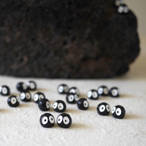 May include: A collection of small, black, round beads with white eye details scattered on a light-colored surface. A large, dark, textured rock is in the background. The beads resemble soot sprites, a whimsical and artistic theme.
