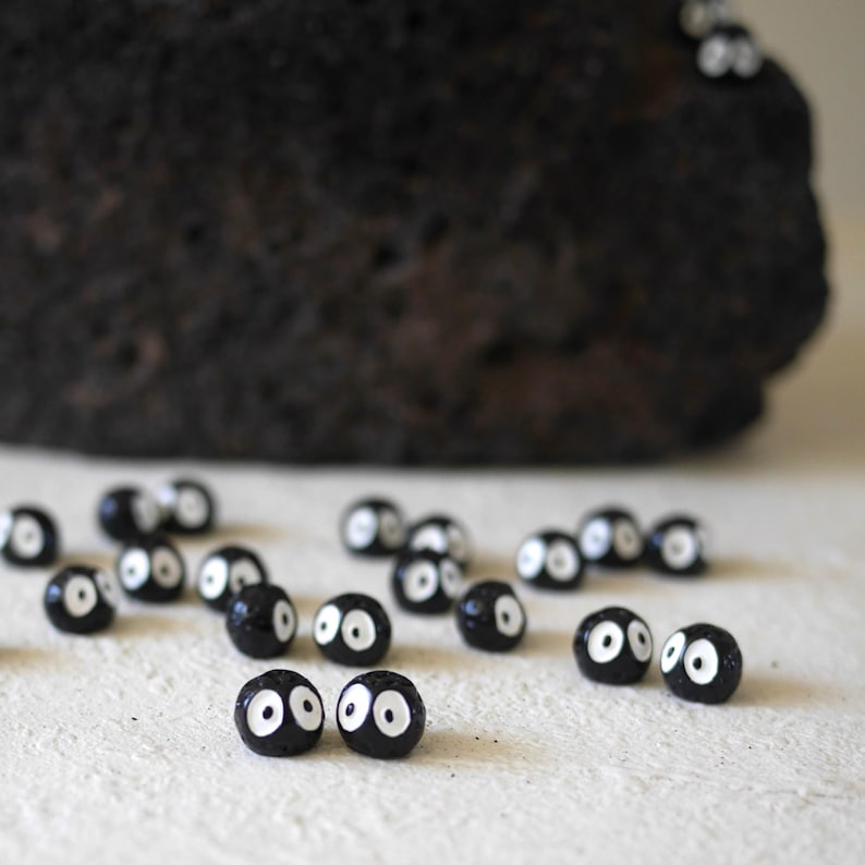 May include: A collection of small, black, round beads with white eye details scattered on a light-colored surface. A large, dark, textured rock is in the background. The beads resemble soot sprites, a whimsical and artistic theme.