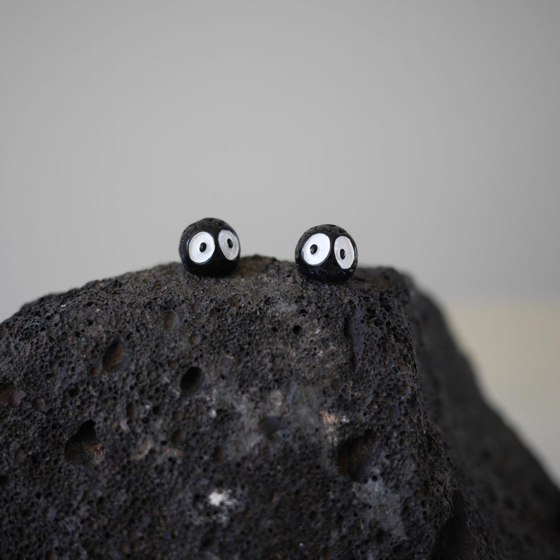 May include: Two small, round, black objects with white oval eyes sit atop a dark, textured rock. The objects resemble cartoon characters, with a simple, minimalist design. The background is a plain, light gray.