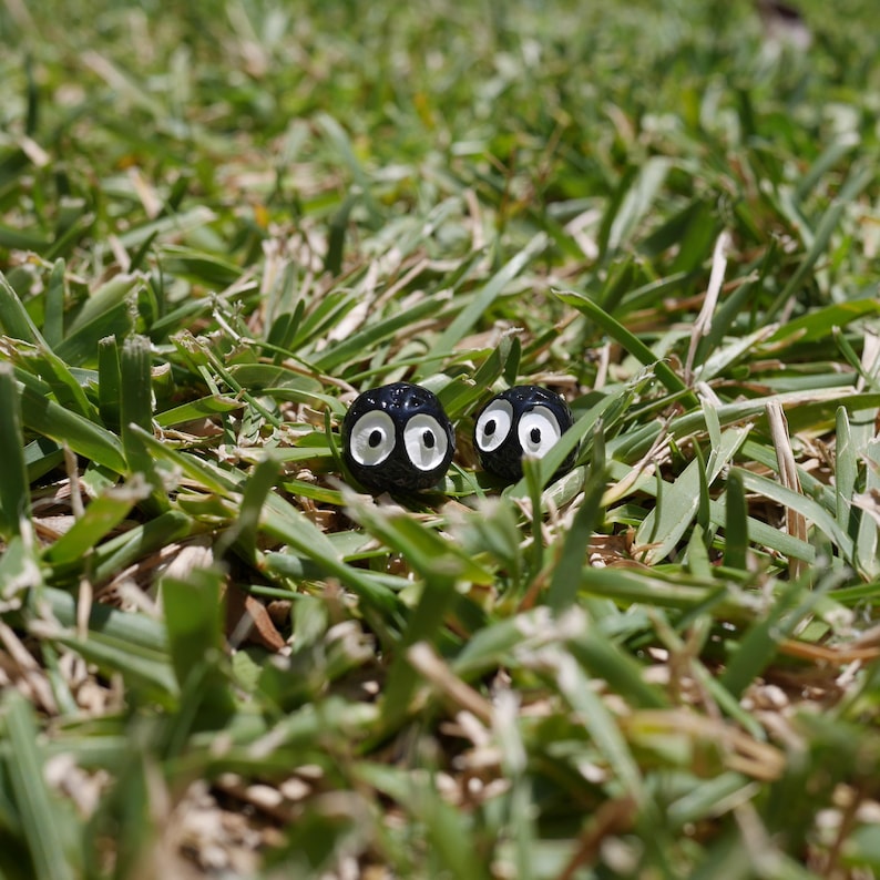 May include: Two small, black, round objects with white eye details are nestled in green grass. The objects have a cartoonish appearance, resembling eyes peeking out from the grass. The scene is set outdoors.