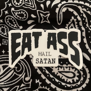 May include: White sticker with black text that reads "EAT ASS HAIL SATAN". The sticker is on a black and white paisley bandana.