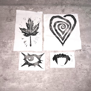 May include: Four black and white linocut prints on white paper. The prints include a maple leaf, a heart with a spiral design, an eye, and a set of vampire fangs. The prints are arranged on a gray surface.