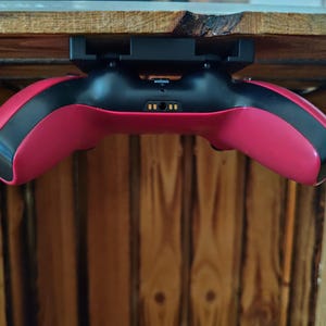 May include: A red and black games controller is mounted under a wooden surface. The controller has a sleek design with a black centre and red sides. The controller is designed for gaming.