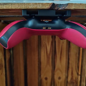 Könnte beinhalten: Ein rot-schwarzer Videospiel-Controller, der in einer schwarzen Kunststoffhalterung montiert ist. Der Controller verfügt über einen zentralen Ladeanschluss mit sichtbaren goldenen Kontaktstiften und ist horizontal unter einer Holzoberfläche angebracht.