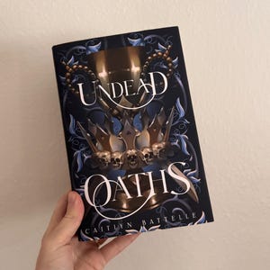 May include: A hardcover book titled "Undead Oaths" with a dark blue cover. The cover features gold and silver illustrations of crowns, skulls, and ornate designs. The author's name, "Caitlyn Battelle," is printed at the bottom.