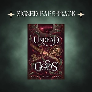 May include: A signed paperback book titled "Undead Gods" by Caitlyn Battelle. The cover features a dark red apple, a skull, a key, and a sword. The background is a dark green with the words "Signed Paperback" at the top.