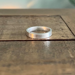 May include: A silver-coloured ring with a flat, brushed metal finish and a simple, circular band design. The ring features a consistent width throughout its circumference, showcasing a clean, unadorned surface with a subtle, matte texture.