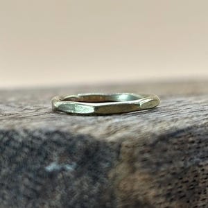 May include: A gold-coloured ring with a faceted design. The ring is set against a rustic wooden surface, creating a contrast between the smooth metal and the textured wood. The ring's shape is geometric.