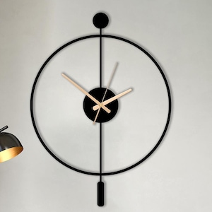 Op de afbeelding: Een moderne zwarte metalen wandklok met een cirkelvormig ontwerp en goudkleurige wijzers hangt aan een witte muur. Twee perzikkleurige stoelen met zwarte poten en een kleine zwarte tafel met een theeset staan ervoor. Een zwarte vloerlamp staat links.