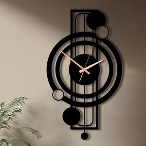 Può includere: Un moderno orologio da parete nero con un design geometrico. L'orologio presenta un quadrante circolare con lancette color oro rosa e una serie di cerchi e forme sovrapposti. L'orologio è montato su una parete di colore neutro, con piante in primo piano.