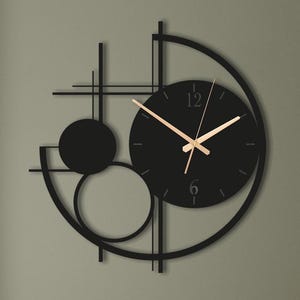 Può includere: Un orologio da parete in metallo nero caratterizzato da un design geometrico asimmetrico con quadrante circolare, lancette sottili color oro e i numeri 12 e 6 in un carattere semplice. La struttura include linee e cerchi che si intersecano.