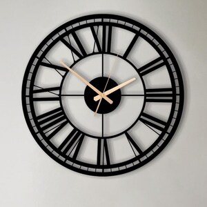 May include: A large circular wall clock featuring a black metal frame with open-work Roman numerals. The clock has a central black circular hub and thin, metallic gold-colored hour and minute hands that contrast against the dark frame.