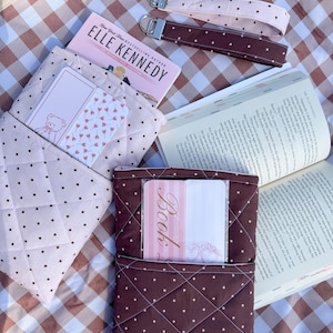 Puede incluir: Dos fundas de tela acolchada para libros en rosa claro y marrón oscuro con estampado de lunares, cada una con marcapáginas. Incluye dos llaveros de muñeca de tela a juego, un libro de Elle Kennedy y un libro abierto con texto visible.