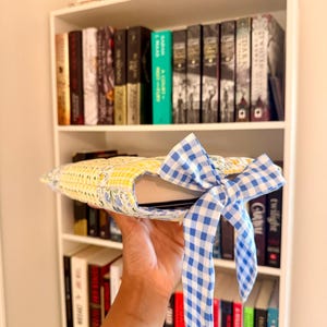May include: A book sleeve with a yellow floral and blue gingham pattern, tied with a blue and white gingham bow. The sleeve is holding a book, with a bookshelf in the background.