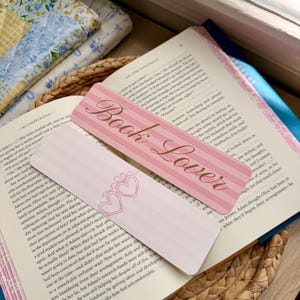 Puede incluir: Dos marcapáginas rosas con las palabras "Book Lover" y diseños de corazones. Los marcapáginas están sobre un libro abierto. La escena está ambientada sobre una superficie de madera con una cesta tejida y tela en el fondo.