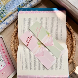 Puede incluir: Dos marcapáginas rectangulares con el texto "In my girlhood era!" y una ilustración de cóctel, apoyados en un libro abierto. Los marcapáginas son rosas y verdes. El libro está en una cesta tejida.